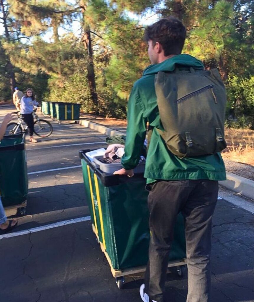 A student seen from behind wearing a green jacket and backpack, pushing a wheeled bin on dorm room move in day.