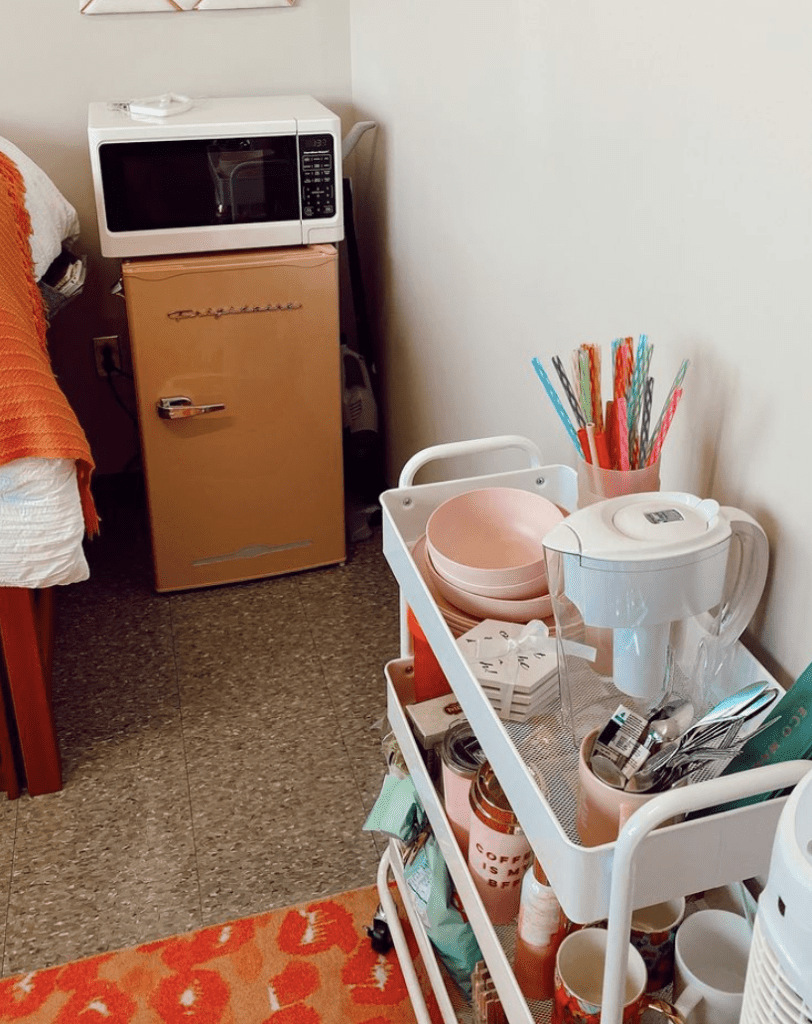 A compact dorm room kitchenette area with a vintage-style orange mini-fridge and a microwave on top. A white utility cart is stocked with kitchen essentials like bowls, a water filter pitcher, and colorful utensils.