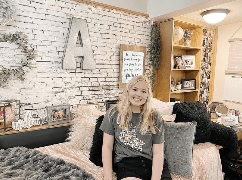 A smiling young woman sitting on a bed in a dorm room with a faux brick wallpaper accent wall. A large metal letter 'A' hangs above the bed, surrounded by inspirational quotes and personal photos.