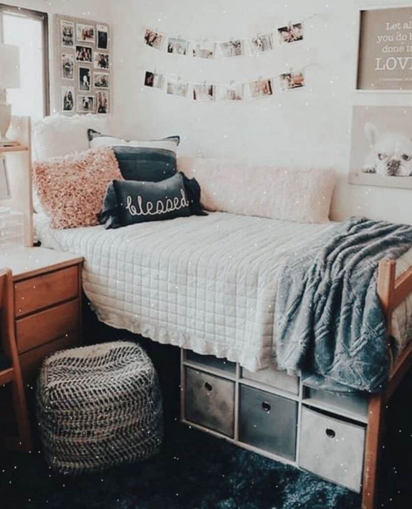 A snug and stylish dorm room bed with a variety of textured pillows, including one with the word 'blessed.' The bed is covered with a checkered comforter and a gray throw blanket. Above the bed, string lights drape over photo collages, and motivational posters add character to the space. Underneath the bed, storage cubes provide an organized solution for personal items