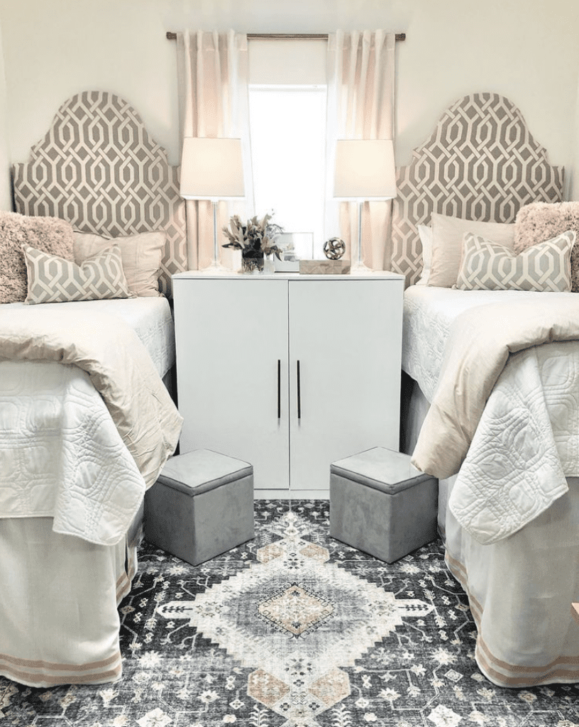 Symmetrically designed dorm room with two twin beds featuring patterned upholstered headboards and neutral bedding. Between the beds is a window with sheer pink curtains, flanked by matching white bedside tables with lamps. In front of the beds are two gray ottomans on a decorative area rug with a geometric pattern.