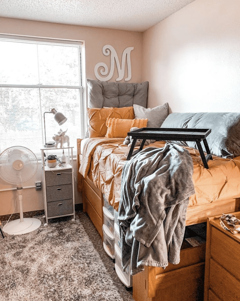 A dorm room featuring a single bed with mustard yellow bedding and a large padded gray headboard against a neutral wall with an ornamental letter 'M.' A laptop tray, a shaggy area rug, and a bedside table with a lamp and small decorative items create a functional yet cozy student living space.