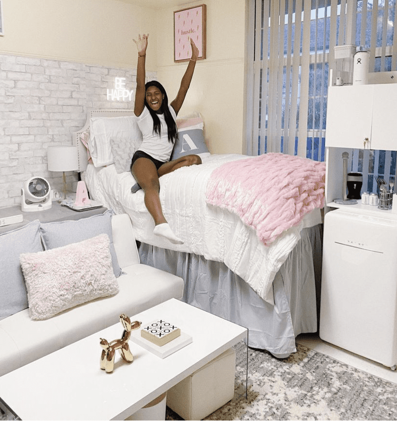 A joyful young woman sits on a bed with white and pink bedding in a well-decorated dorm room. The room features a faux brick wall, inspirational wall art, and a cozy futon with throw pillows.