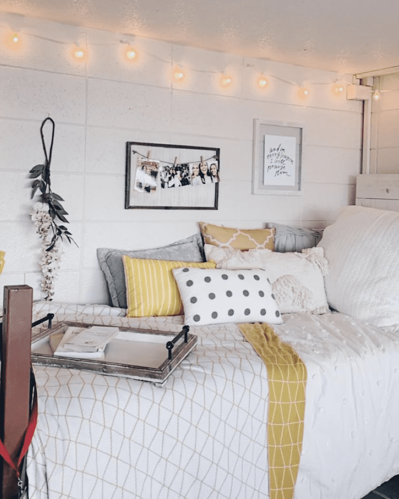 Inviting dorm room with a bed covered in white and geometric-patterned bedding, accented by yellow and gray pillows. Above the bed, string lights add a warm ambiance, next to framed pictures including a group photo and a quote print.