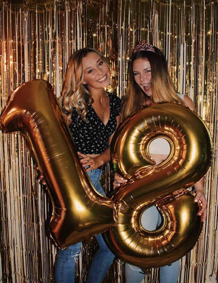 Two smiling young women celebrating, standing in front of a shimmering gold foil backdrop. They are holding large metallic number balloons that read '18'.