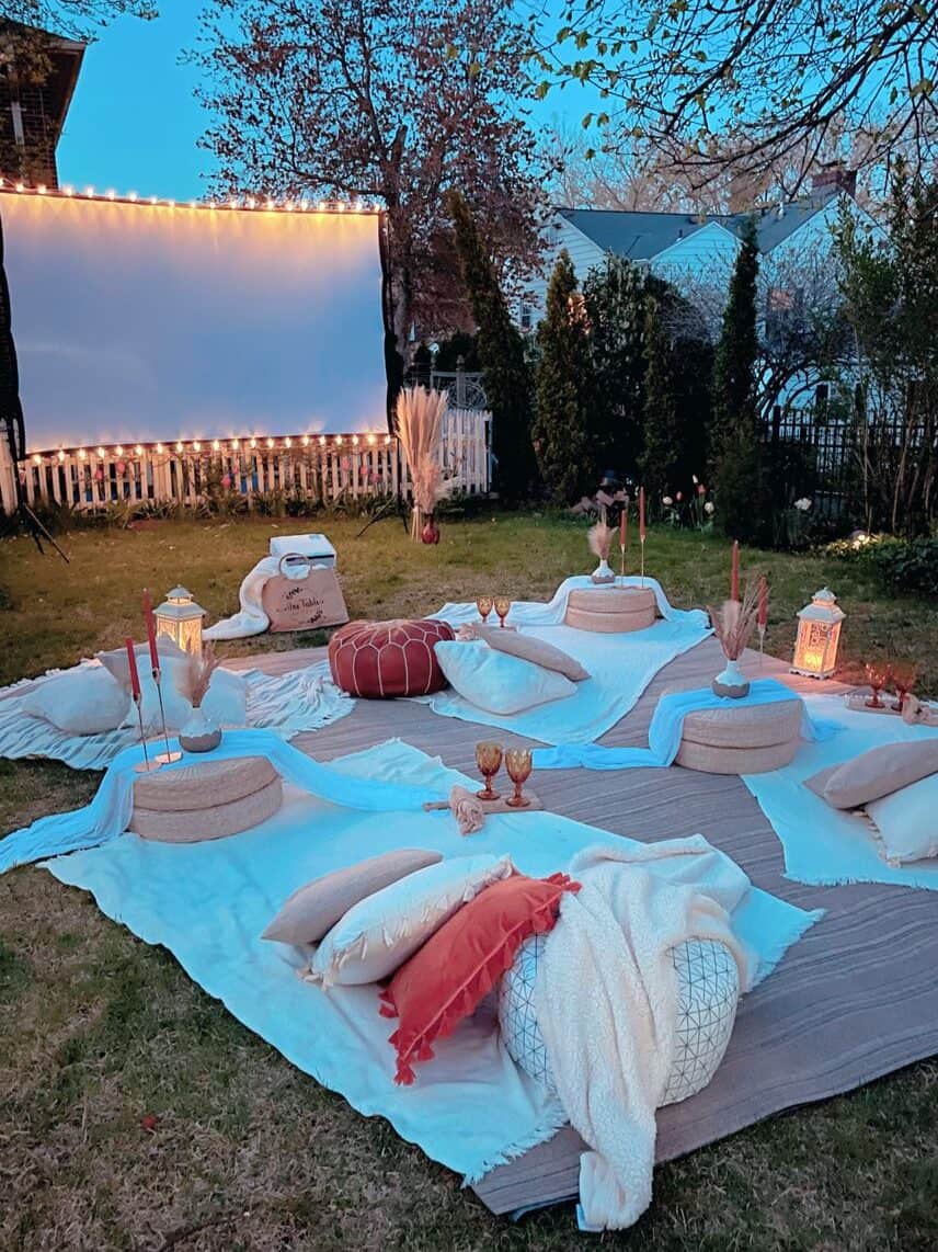 A cozy outdoor movie night setup in a backyard during dusk. The scene includes a large projector screen, string lights along the fence, floor cushions, blankets, and a lantern on a wooden deck.