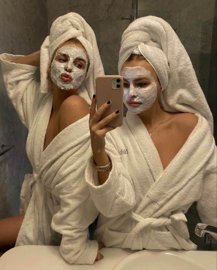 Two individuals in a bathroom, sporting white facial masks and wrapped in matching bathrobes with their hair up in towels, take a mirror selfie with a smartphone.