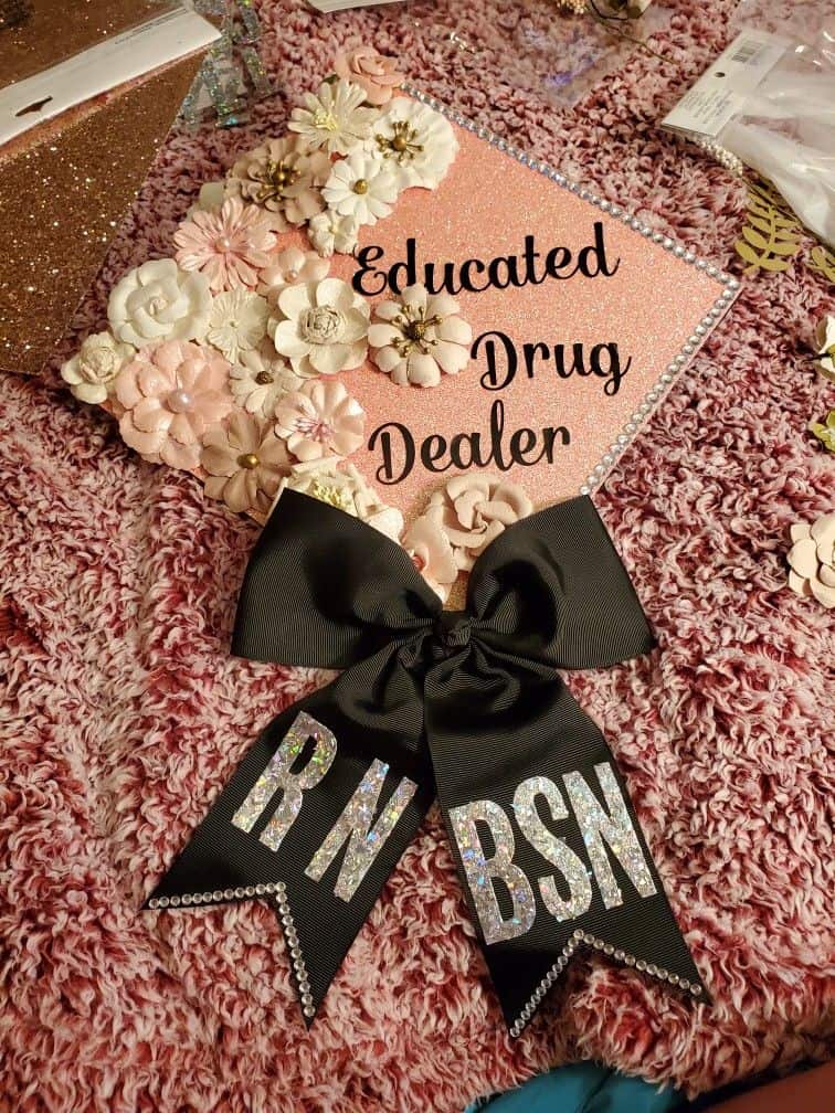 A graduation cap with a humorous twist, featuring the phrase 'Educated Drug Dealer' in black cursive script on a glittery coral background. The cap is adorned with a variety of cream and pink paper flowers along the edge and a large black bow with the letters 'RN BSN' in sparkling silver.