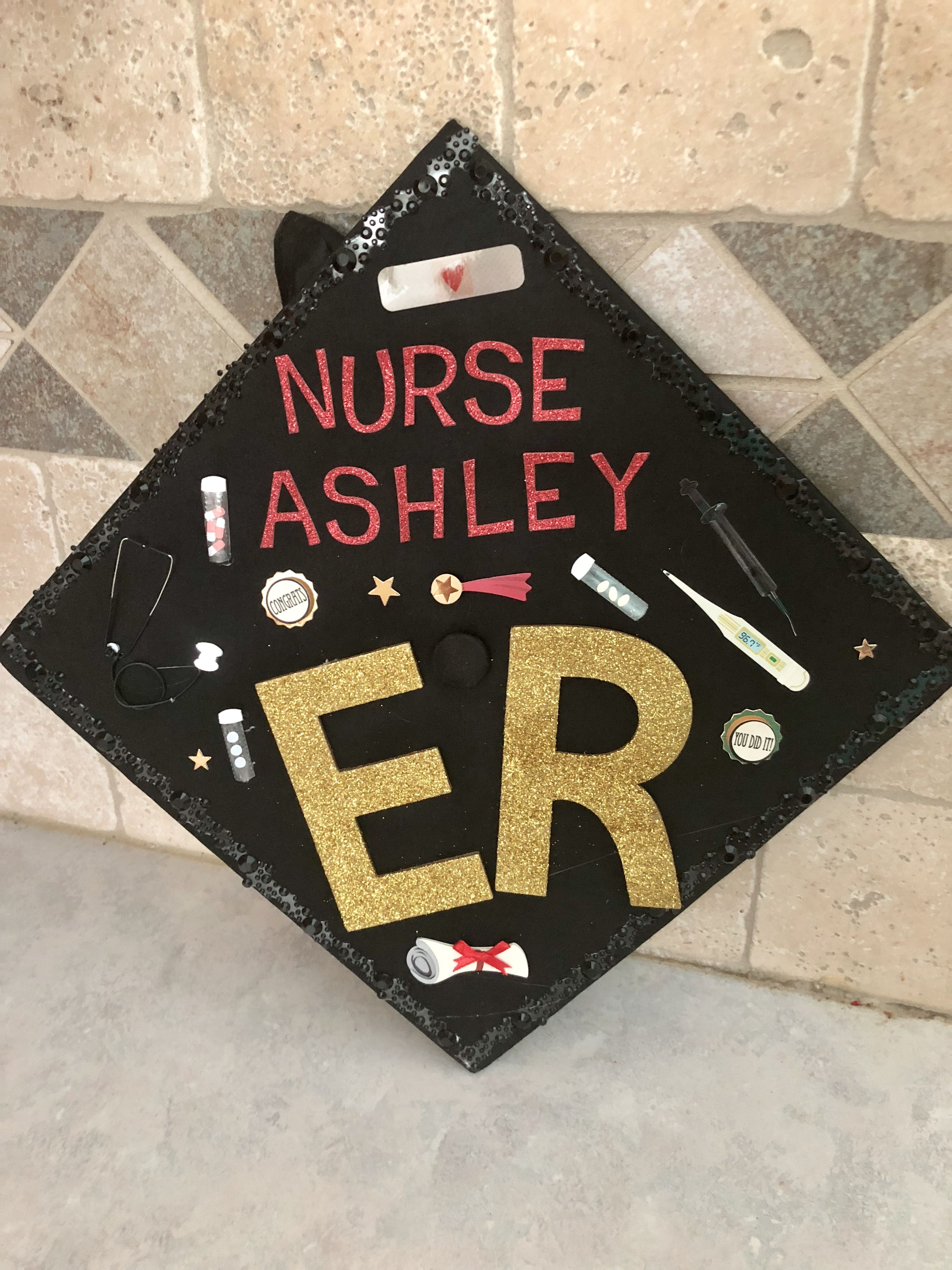 A customized graduation cap with sparkling black sequins, featuring the text 'NURSE ASHLEY ER' in bold red and gold letters. The cap is adorned with themed decorations including a stethoscope, syringe, pill bottle, bandage, and a 'You did it!' badge.