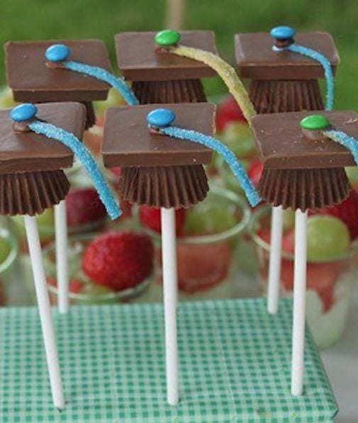 The image features a creative graduation-themed dessert presentation. Mini chocolate candy squares are mounted on white lollipop sticks, resembling graduation caps. A thin, colorful sour candy strip is attached to the top as a tassel, and a single round M&M represents the button of the cap.