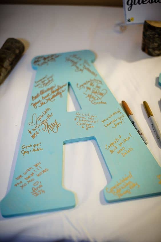 A large wooden letter 'A' painted in light blue, covered with heartfelt handwritten messages in gold and silver ink, part of a guest signing station at a graduation party.