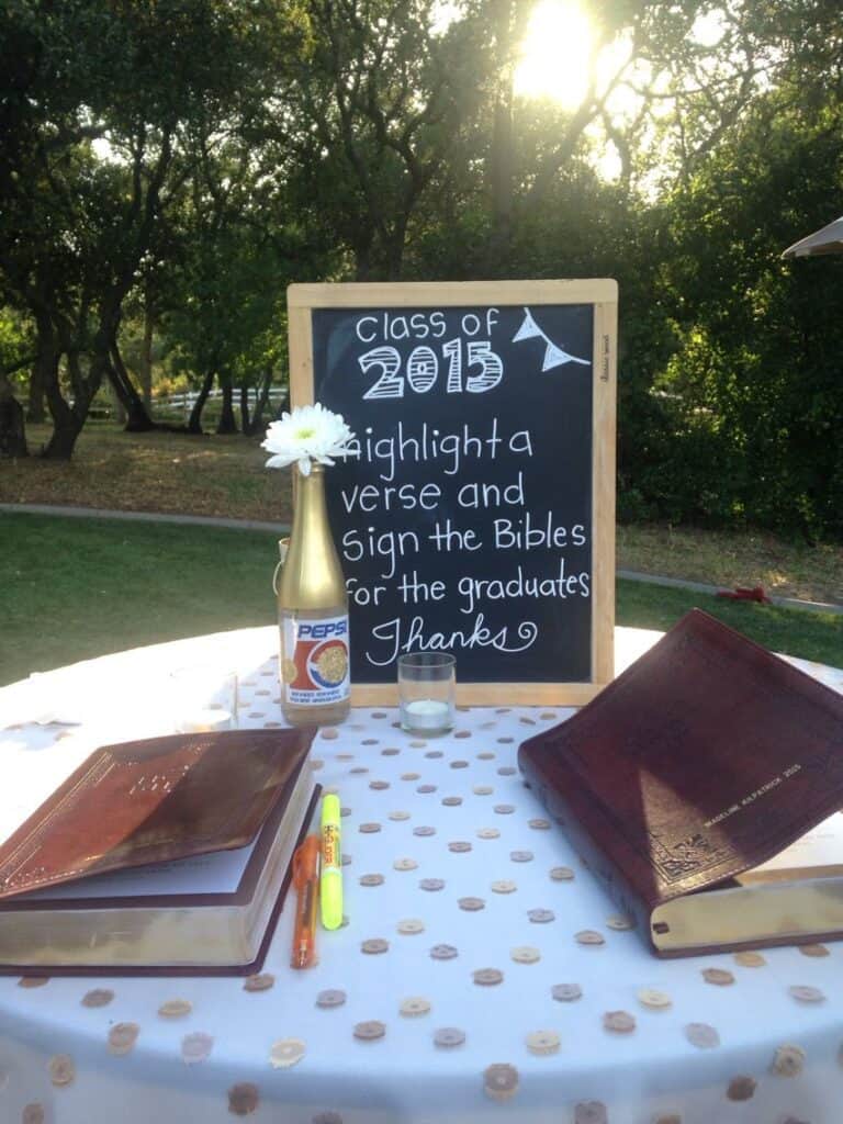 An outdoor graduation event setting with a chalkboard instructing guests to highlight a verse and sign the Bibles, accompanied by a highlighter and pen.