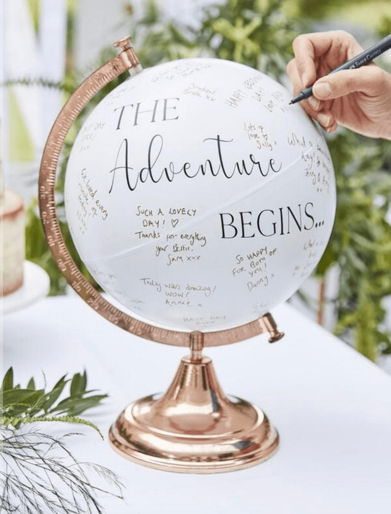 A hand signing a white globe with a rose gold stand and the inscription 'THE ADVENTURE BEGINS...' surrounded by handwritten well-wishes and signatures, used as a creative graduation party guest book alternative.