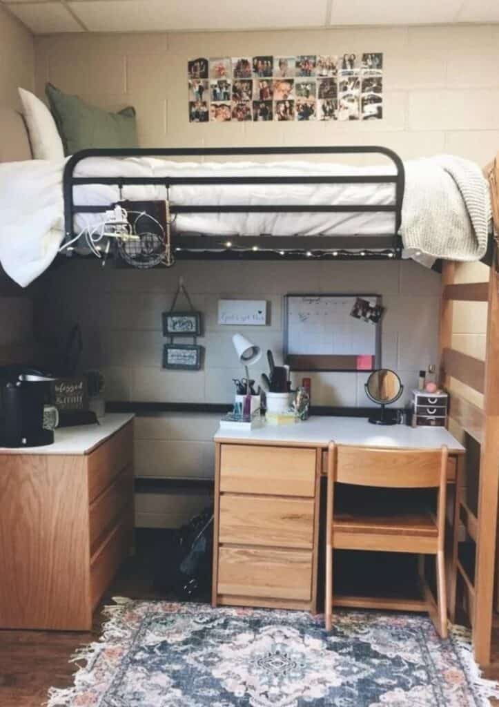 A space-efficient dorm room with a lofted bed and a cozy study area underneath. The bed is accessorized with white bedding and a knitted throw. Underneath, the desk area includes a wooden chair, storage drawers, and a whiteboard.