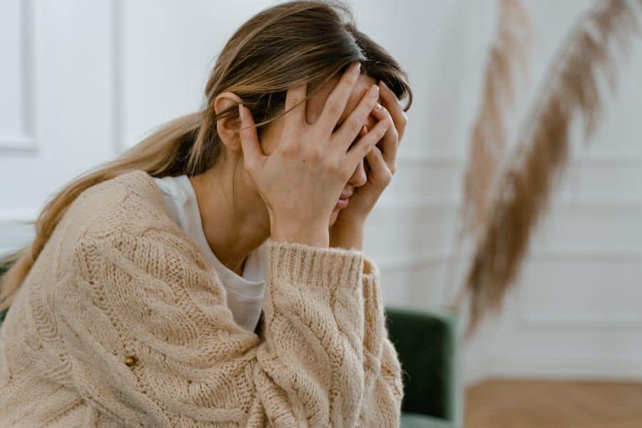 A person sitting indoors with their face covered by their hands in a gesture of stress or exhaustion, wearing a cozy beige sweater, suggesting a moment of overwhelm or concern, possibly related to academic or personal challenges.