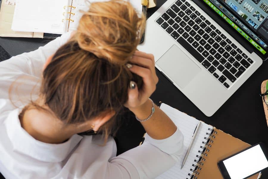 An overhead view of a person at a desk, appearing stressed with one hand on their head, surrounded by a laptop displaying an editing program, notebooks, and a smartphone, indicative of a busy work or study environment.