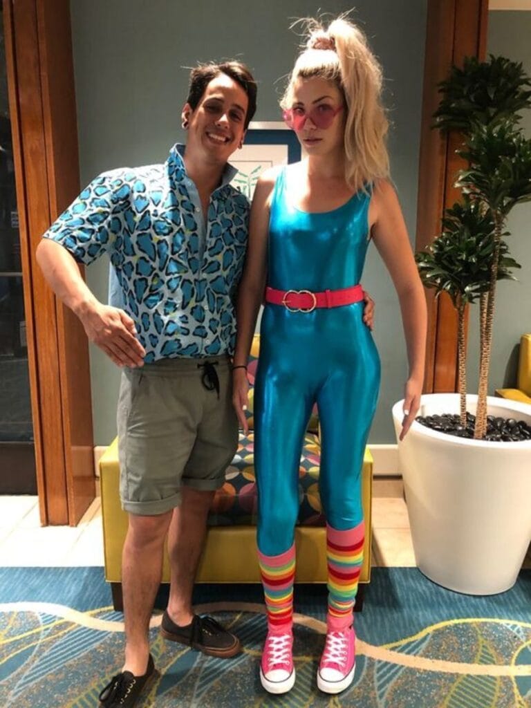 A man and a woman in vibrant costumes posing for a picture, with the man wearing a blue patterned shirt and shorts, and the blonde woman in a shiny turquoise jumpsuit, pink belt, striped knee-high socks, pink sneakers, and pink sunglasses, dressed as Barbie