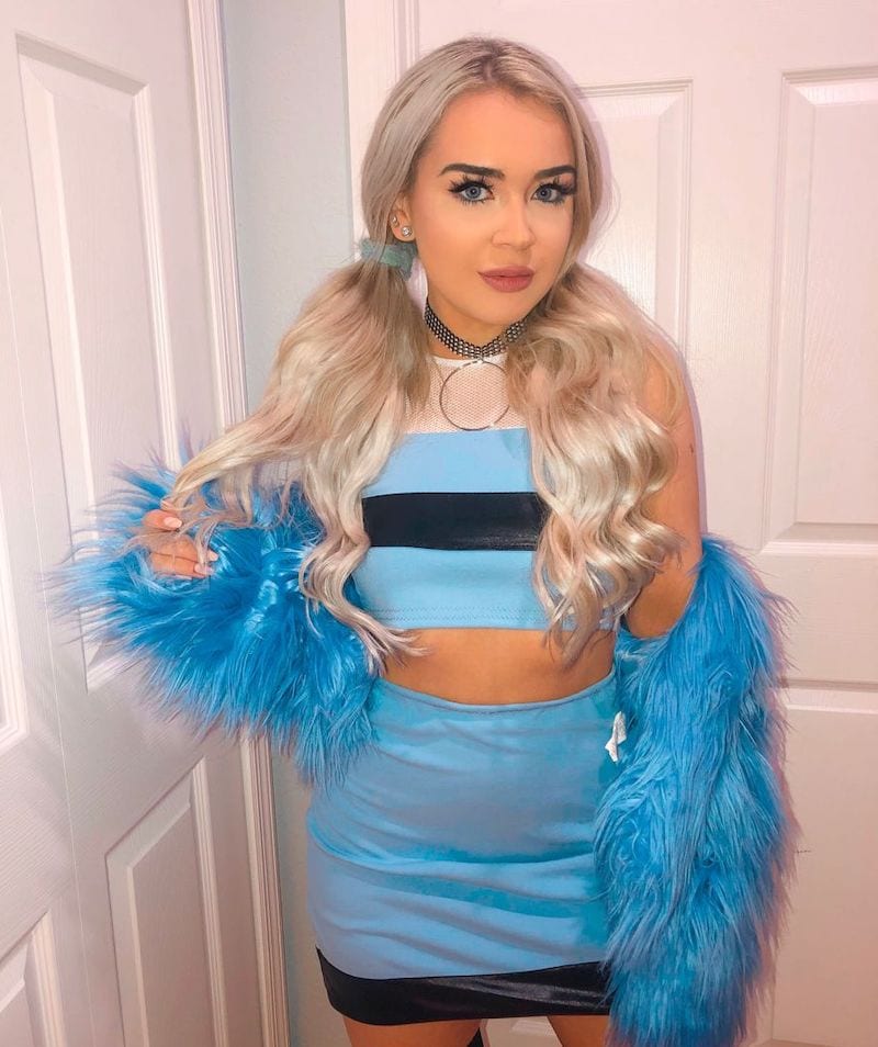 A person with blonde hair posing in a Bubbles Halloween costume, featuring a blue cropped tank top and skirt, accented with a fluffy blue jacket and a choker necklace