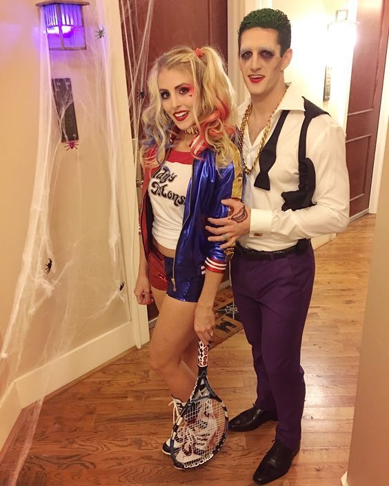 Two people in Halloween costumes, with one person dressed in a Harley Quinn costume, featuring a red and blue jacket, white 'Daddy's Lil Monster' T-shirt, and red and blue shorts, and the other person in a Joker-themed costume with white face makeup, green hair, purple pants, and a white shirt with suspenders, standing in a hallway decorated with Halloween cobwebs.