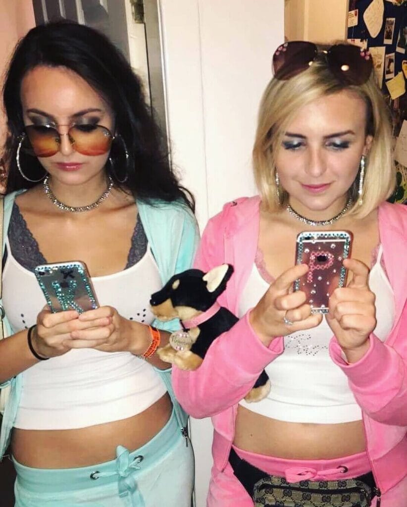 Two women dressed in early 2000s fashion with crop tops, low-rise bottoms, and oversized sunglasses, are focused on their bedazzled flip phones. One is holding a small toy dog, contributing to the retro theme.