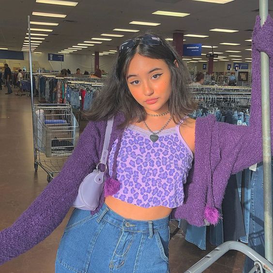 A young woman stands in a thrift store, striking a pose with a shopping cart. She sports a Y2K fashion ensemble featuring a purple leopard print crop top, a chunky choker necklace, and a cozy, purple cardigan with fun pom-pom details. 