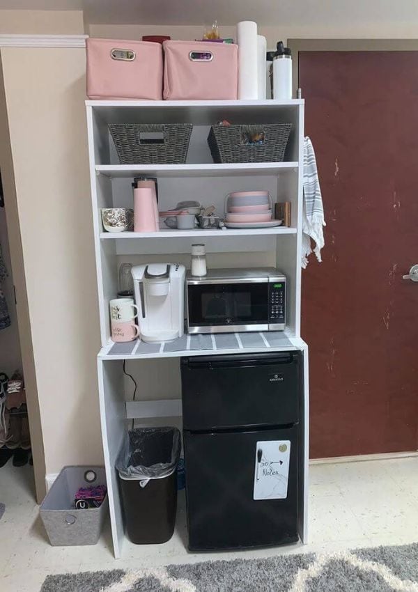 A practical dorm room kitchen setup with a black mini-fridge and freezer, topped with a microwave. A white shelving unit beside it holds kitchen supplies.