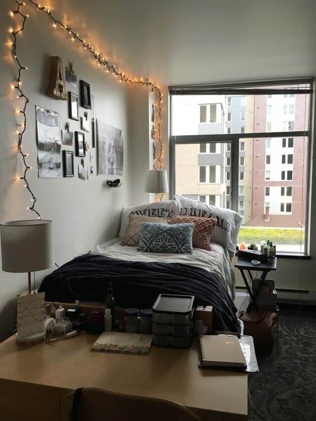 A cozy dorm room with a large window offering a view of the campus. The room is decorated with string lights and a collage of wall art above the bed, which is made with a navy blue comforter and decorative pillows. A desk at the foot of the bed is scattered with notebooks, containers, and a lamp, creating a lived-in and personalized atmosphere for study and relaxation.