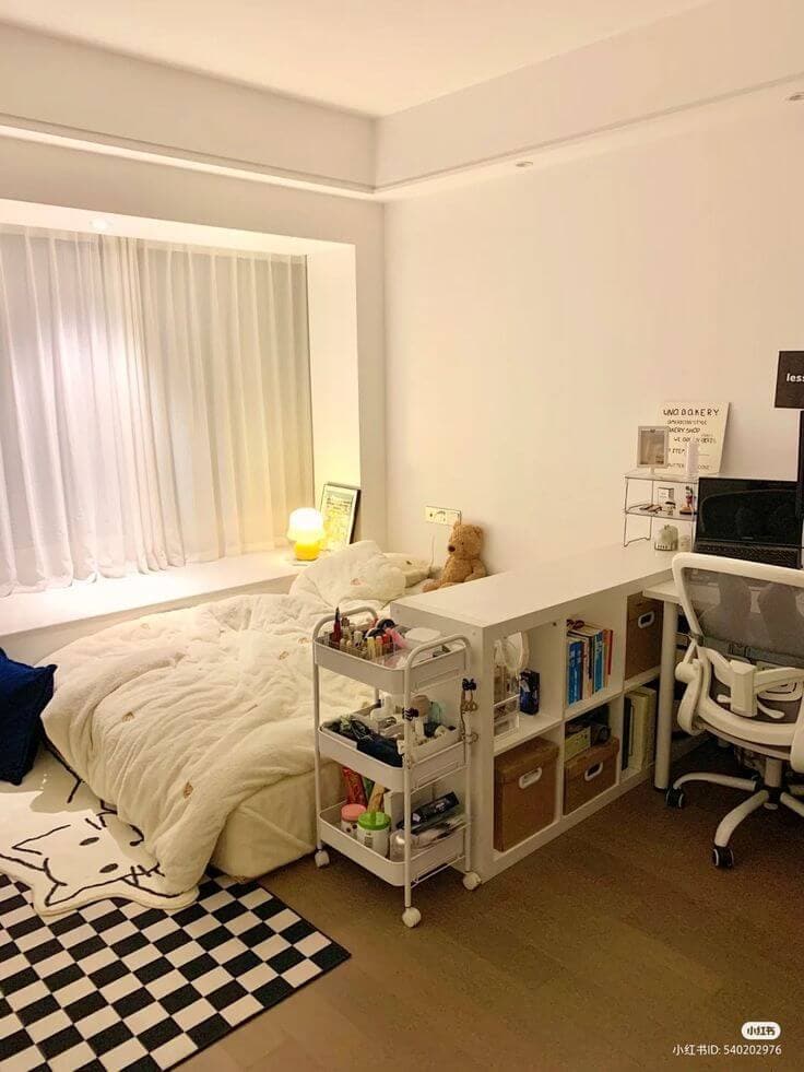 A minimalist dorm room with a single bed covered in a white comforter and a plush toy on it. Beside the bed is a utility cart with various personal care items and a study desk with a laptop and notes.