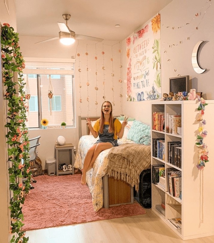 A bright and cheerful dorm room with a young woman sitting on a bed covered in cozy bedding with floral and aquatic hues. The room is decorated with string lights, wall art featuring motivational quotes, and green vines along the window.