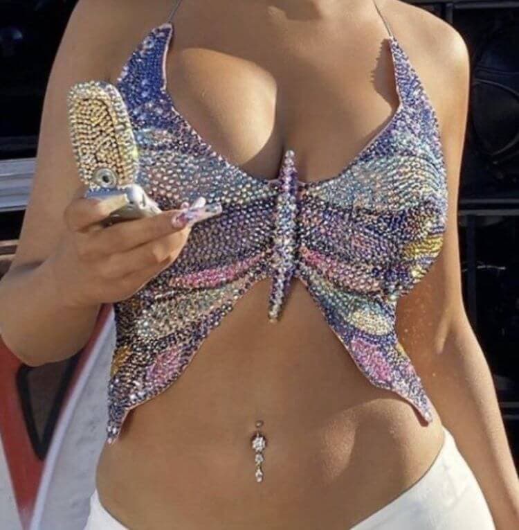 Close-up of a woman's torso wearing a Y2K-inspired butterfly crop top adorned with multicolored rhinestones, creating a dazzling effect. She holds a flip phone with a matching bedazzled case, and her belly button is accentuated with a drop pendant.