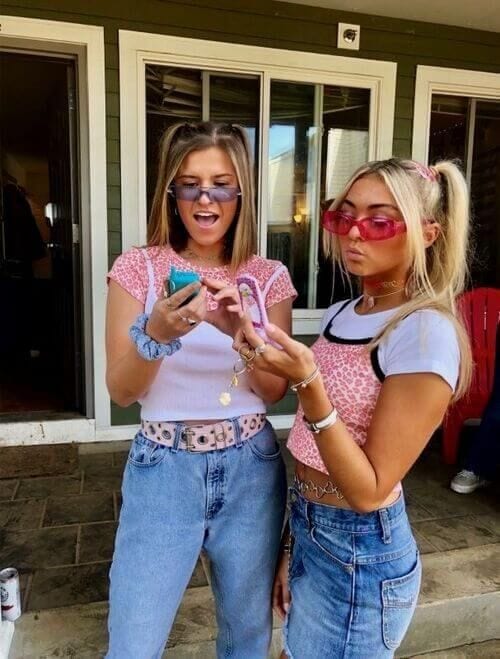 Two young women stand outside, showcasing their Y2K-themed outfits. One wears a pink crop top, high-waisted jeans, and blue-tinted sunglasses, while the other sports a white tee under a pink patterned crop top with matching pink sunglasses and high ponytails. Both accessorize with chunky belts and are engaged with a phone and making hand gestures.