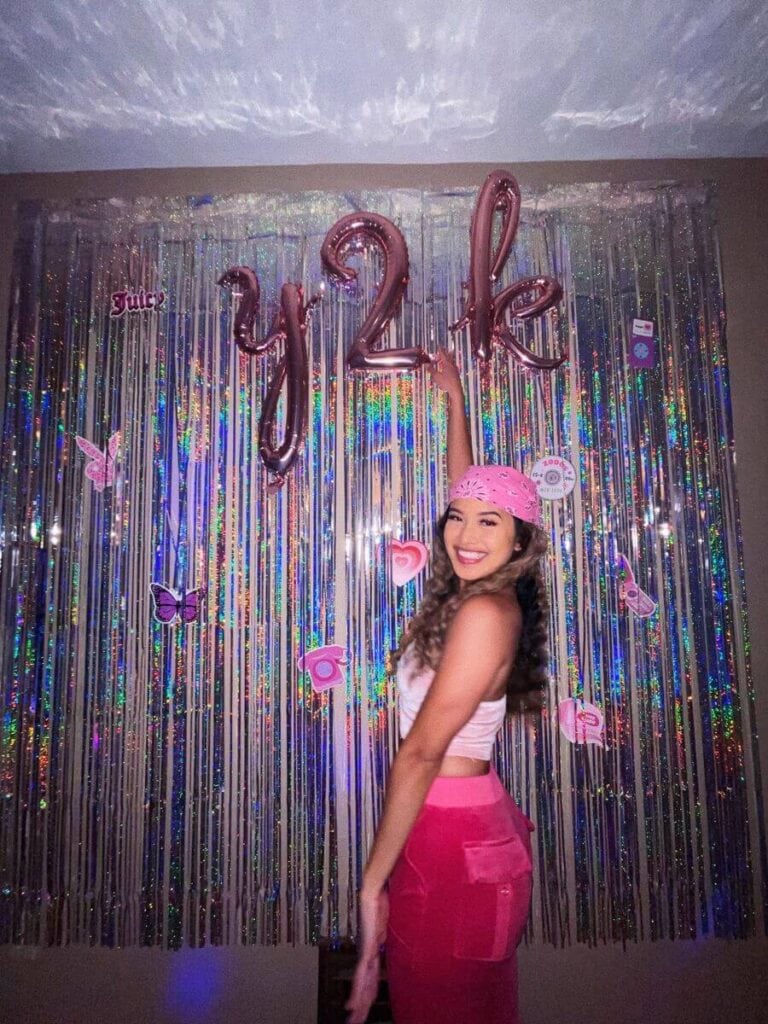 A joyful woman at a Y2K-themed party, wearing a pink bandana, white tank top, and pink cargo pants, stands in front of a shiny, multicolored tinsel backdrop. Above her, balloons spell 'Y2K' and nostalgic decorations like a 'Juicy' logo and retro icons adorn the scene.