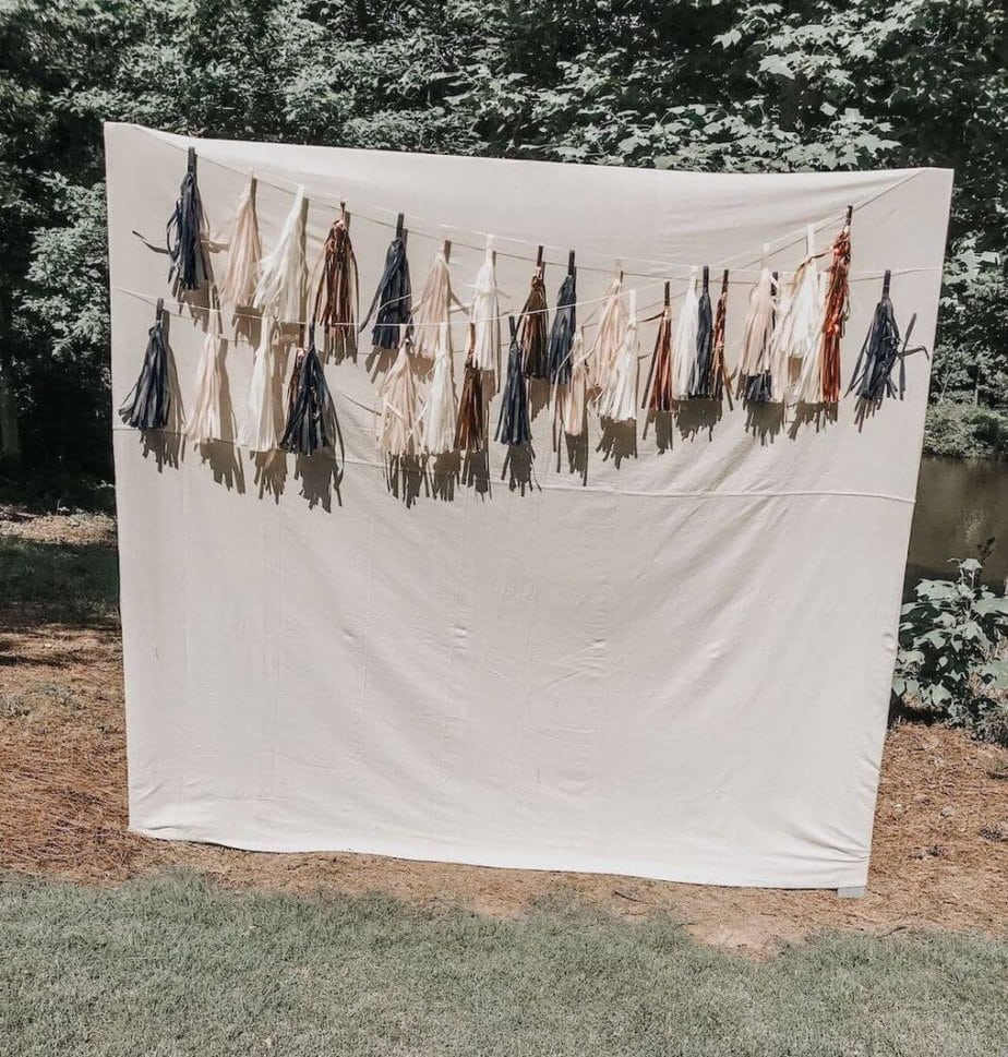 A simple outdoor photo booth backdrop with a white sheet and a garland of tassels in shades of blue, gold, and cream strung across