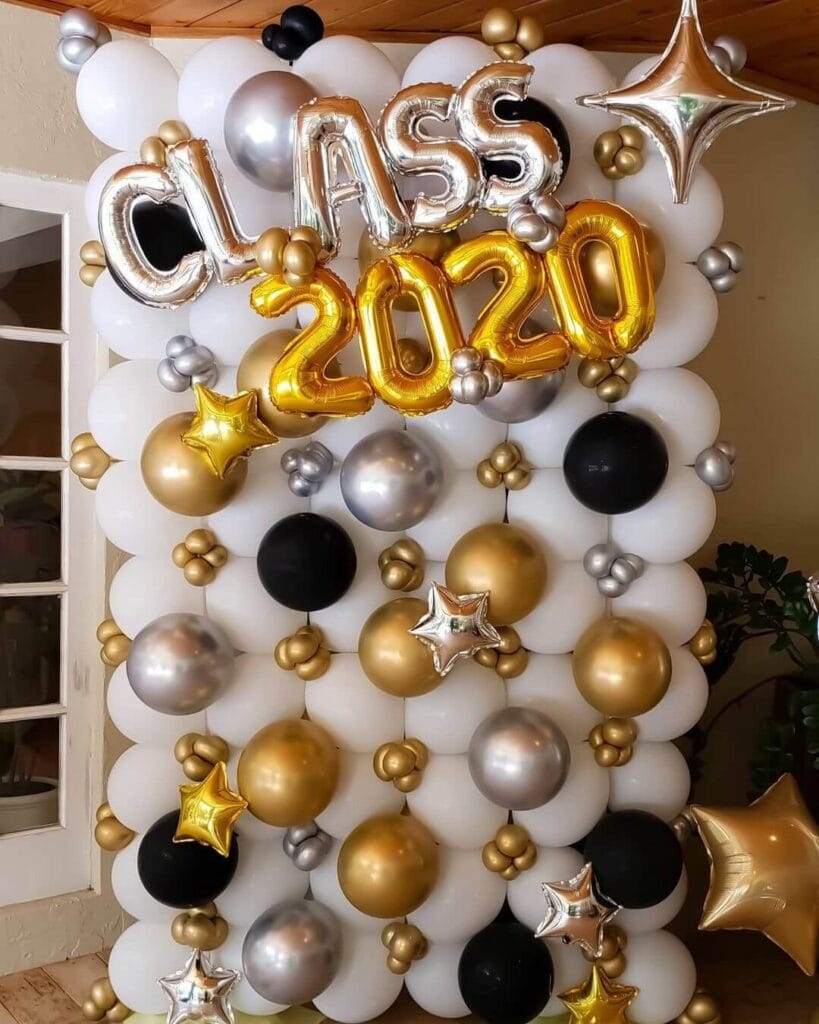 A vibrant balloon wall photo booth at a graduation party featuring gold, silver, black, and white balloons of varying sizes, including star-shaped balloons, with bold 'CLASS 2020' letter balloons in the center