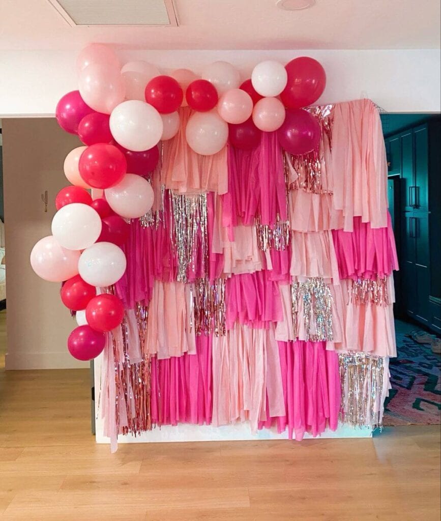 A festive photo booth backdrop featuring a cascade of pink and red balloons on the top with layers of pink fringe and silver metallic tinsel.