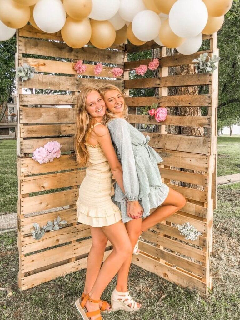 Two young women pose in front of a wooden pallet photo booth decorated with a balloon garland in shades of white and gold, accented with pink flowers, at an outdoor graduation party.