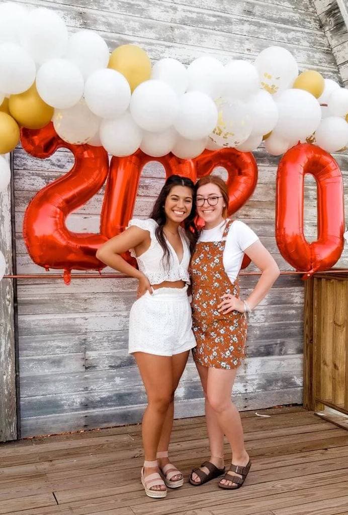 Two smiling friends pose together at a graduation party in front of a weathered wooden backdrop, accented by a balloon arch with the numbers '2020' in bold red.