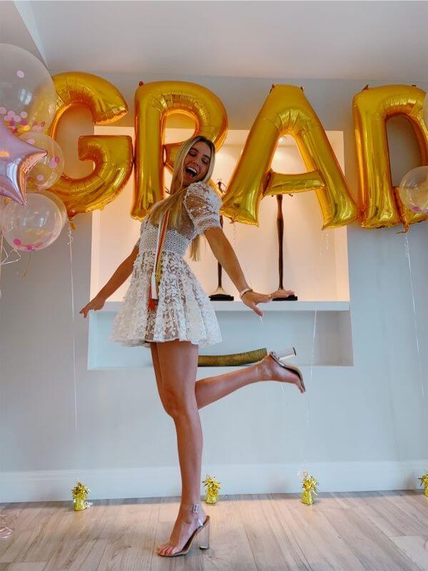 A girl in a white lace dress poses with one foot lifted, smiling in front of a 'GRAD' gold balloon graduation party photo booth backdrop.