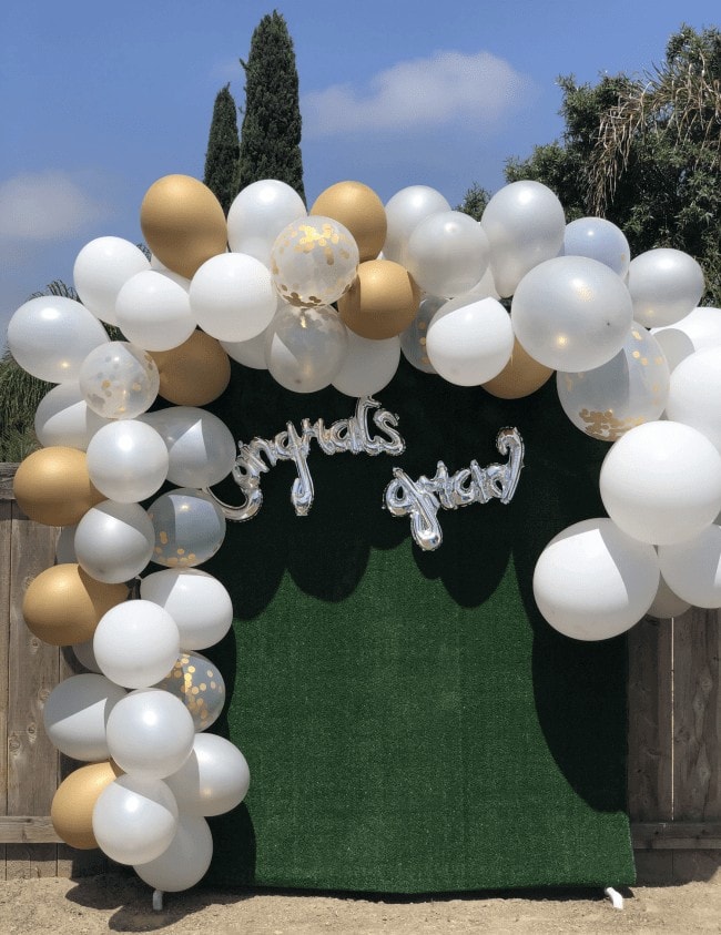 Outdoor graduation photo backdrop featuring a balloon garland in shades of white and gold. The backdrop is adorned with silver 'congrats grad' script balloons, set against a green hedge.