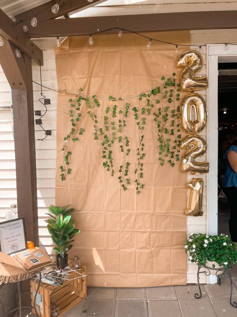 A DIY photo booth backdrop with a brown paper background adorned with hanging green ivy and golden balloons shaped as '2022', set up for a graduation party.