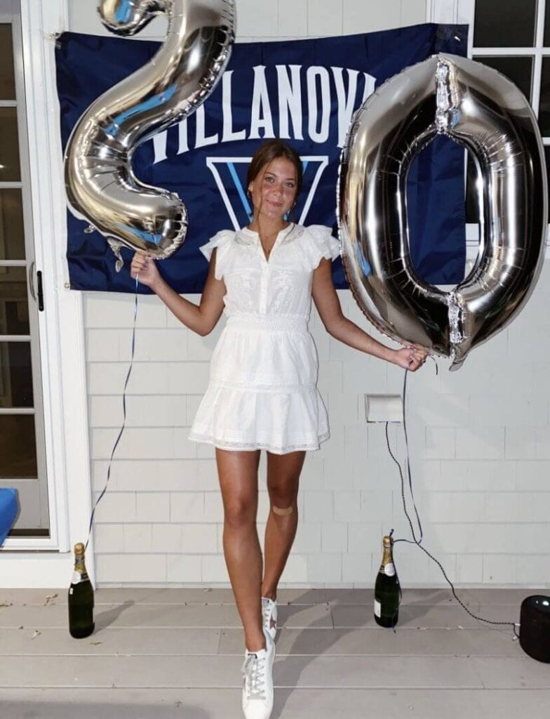 A graduate in a white dress and white sneakers holds up silver number balloons in front of a Villanova University flag