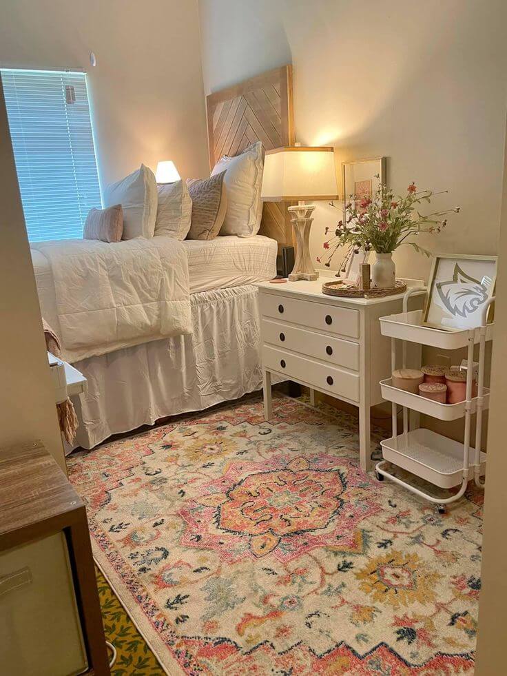 An elegantly decorated dorm room with a raised bed dressed in white linens and multiple pillows. A wooden headboard adds a rustic touch, complemented by a white dresser and nightstand with a classic lamp.