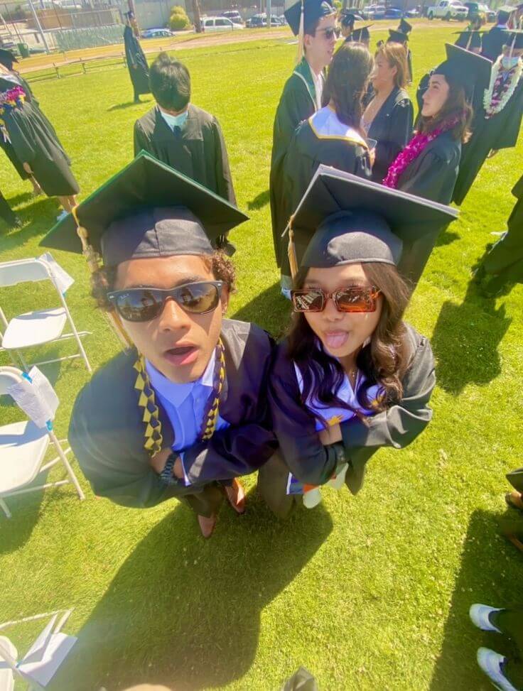 Two excited graduates in black robes and caps make funny faces at the camera, with fellow graduates in the background on a sunny day.