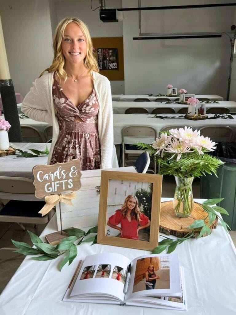 A smiling young woman stands behind a graduation party welcome table featuring a photo frame, a 'Cards & Gifts' sign, and an open photo album, with banquet tables set in the background.