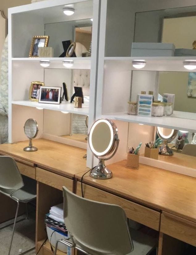 A well-organized dorm desk area with a mirror and built-in shelving. The shelves are adorned with various items including framed photographs, beauty products, and books. Two chairs are placed in front of the desks.