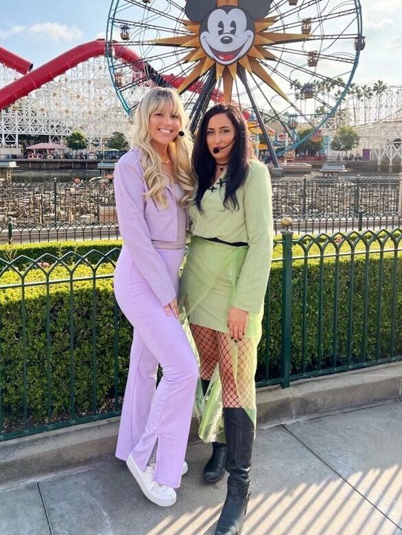 Two individuals posing together at an amusement park with a Ferris wheel in the background; one dressed in a purple tracksuit with blonde hair and white sneakers, the other in a green dress with fishnet tights and black boots, both smiling for the camera.