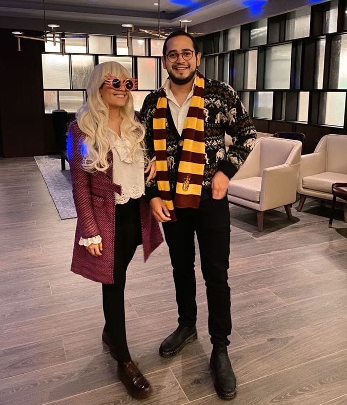 Two people in Halloween costumes posing indoors; one with long blonde hair, round pink sunglasses, and a maroon coat over a white blouse and black pants, the other wearing a Harry Potter-themed scarf, a patterned sweater, and black pants.