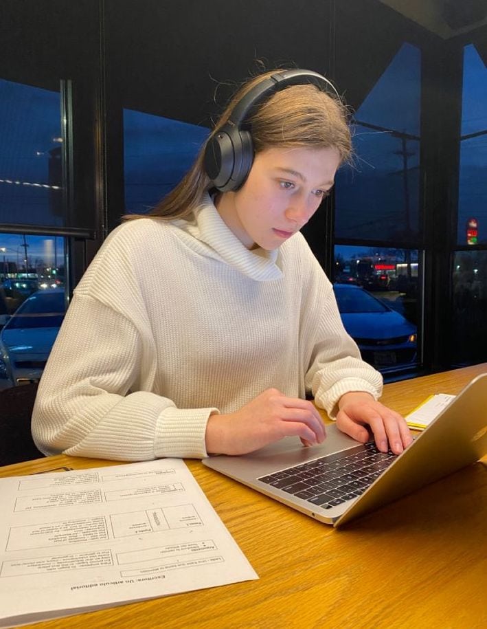A focused student wearing headphones works on a laptop at a table, with study materials in front of her.