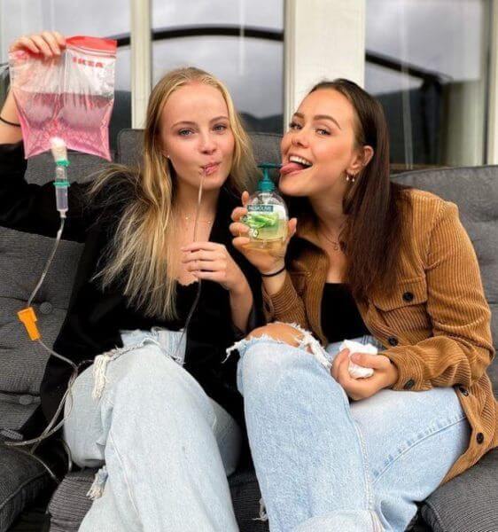 Two young women sitting closely on a couch, playfully participating in an 'Anything But A Cup' party. One is holding a soap dispenser to her mouth like a bottle, while the other pretends to drink from a plastic bag typically used for medical IV fluids.