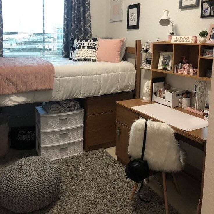 A neatly organized dorm room corner with a single lofted bed covered in pink bedding. Below the bed, storage bins and a white drawer unit are used to maximize space. A study desk with a wooden chair covered in a white faux fur seat cover, a desk lamp, and stationery items is placed beside the bed.